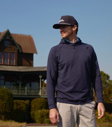 A man wearing white cream pants and a blue hoodie with a hat, standing on what appears to be a golf course with a house and a lush garden in the background.