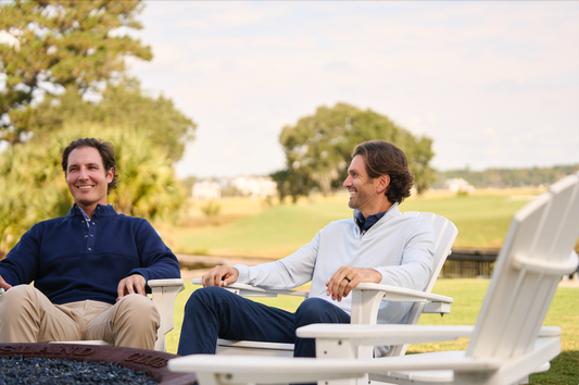 Two golfers in formal attire, smiling and chatting, seated outdoors overlooking a scenic golf course.