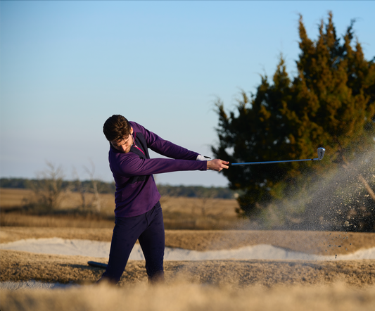 A professional golfer wearing The Traveler Herringbone on a golf course, mid-swing with a golf club. 