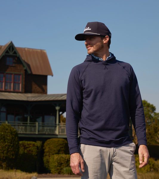 A man wearing white cream pants and a blue hoodie with a hat, standing on what appears to be a golf course with a house and a lush garden in the background.