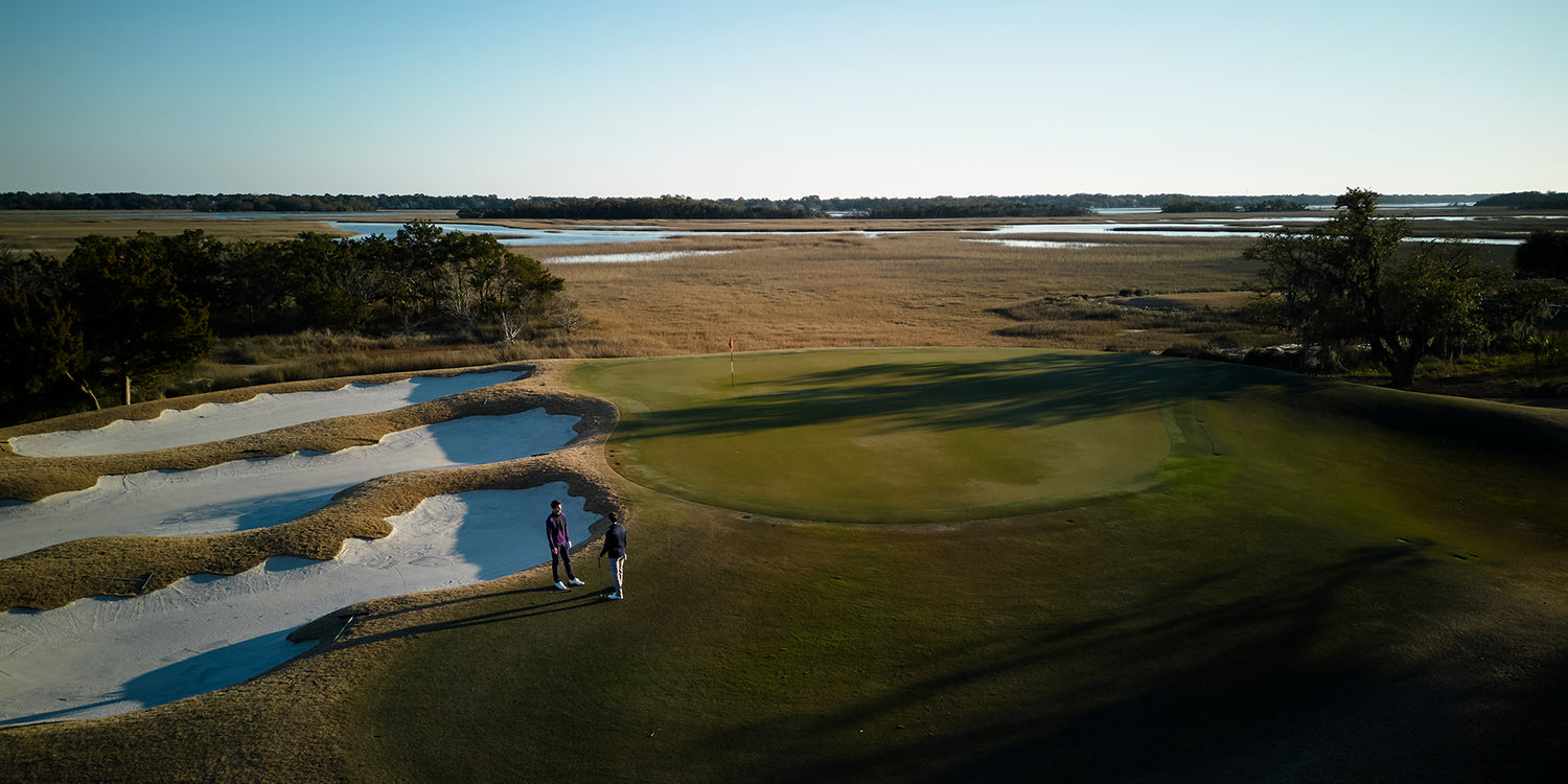 Wide shot of a golf course with polished grass and a hole in the center, two men talking nearby, and a large field beyond the trees in the background.